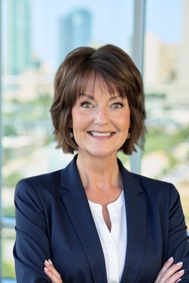 Headshot of a smiling woman with short brown hair, wearing a navy blazer and white shirt, with a blurred office background.